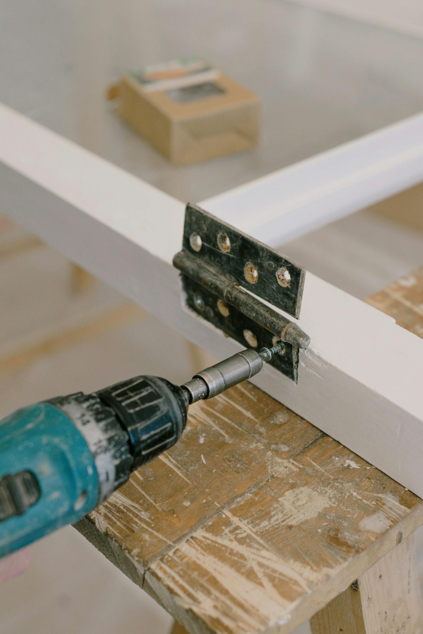 Electric screwdriver with drill attached to metal hinge on painted window placed on wooden workbench in room during relocation works